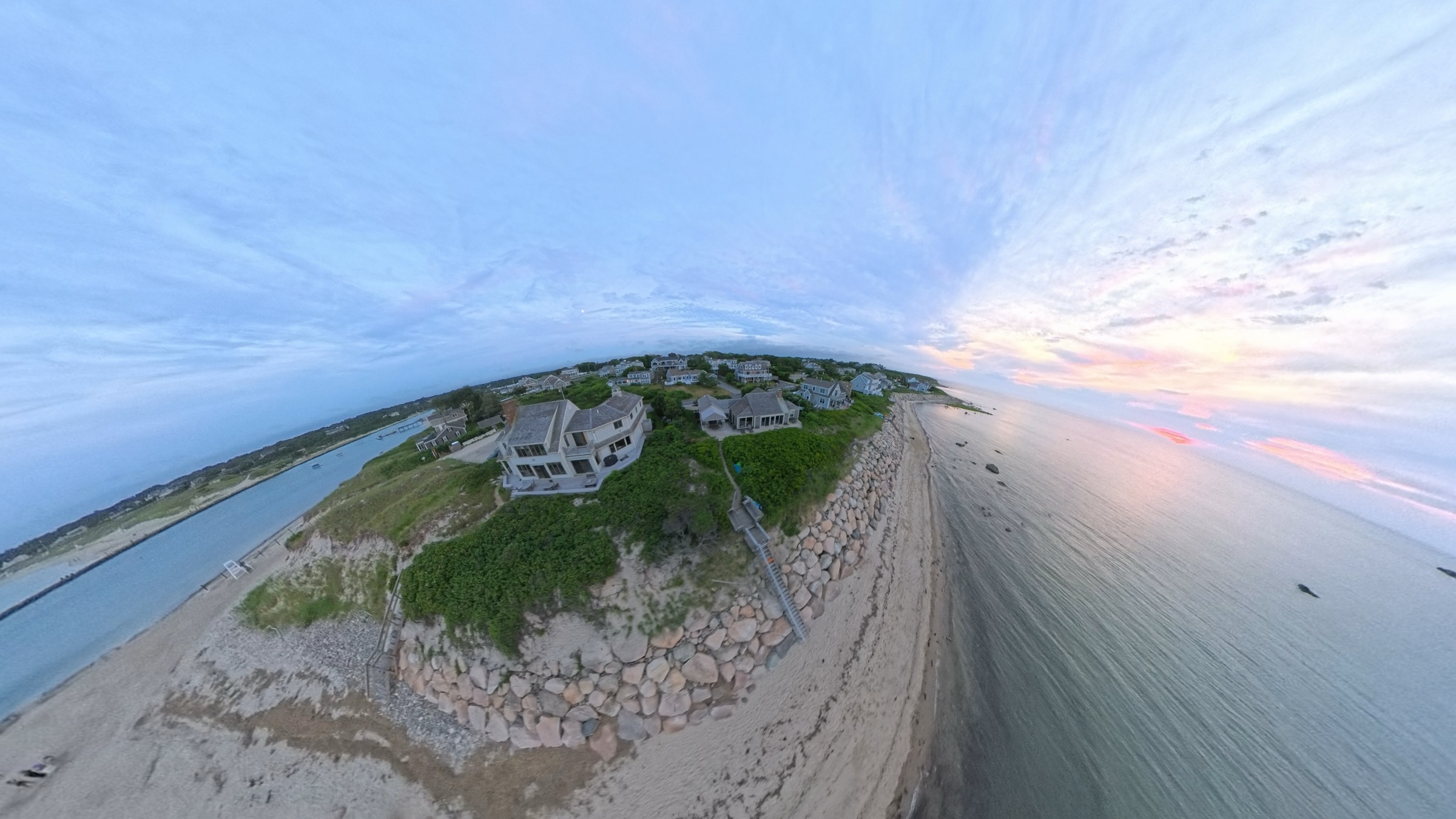 Aerial sunset over Sesuit Harbor in East Dennis, Massachusetts — shingled Cape homes on the bluff above the seawall and beach, with the harbor jetty and inlet to the west.