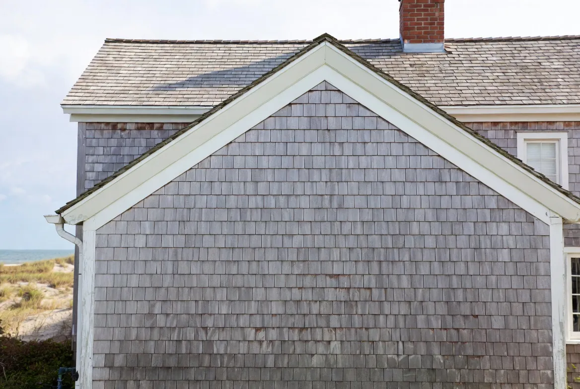Weathered silver-gray cedar shingle siding on a Cape Cod gable end with white trim and a brick chimney, dune grass and the bay behind