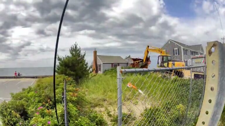 Beachfront home teardown in progress in Dennis Port, MA, with an excavator demolishing the house and the ocean behind