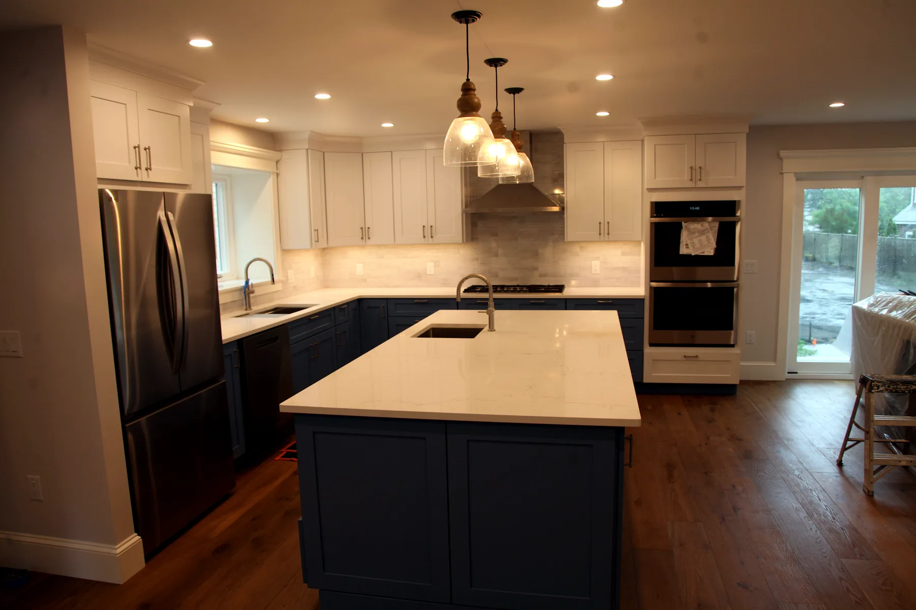 Wide view of finished Cape Cod kitchen with island, built-in oven, and sliding door to the rear yard